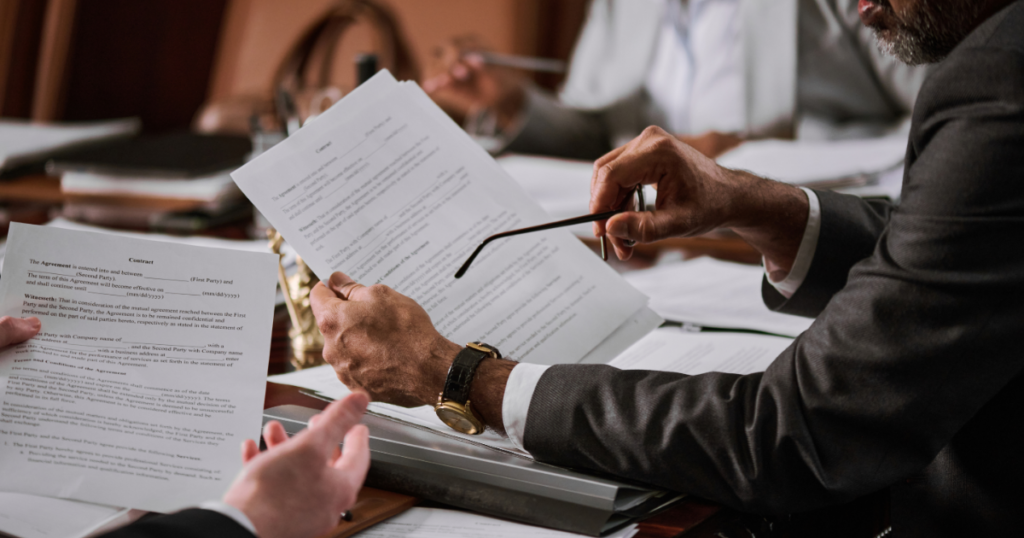 A person in a suit holds a document and glasses while reviewing papers with others in a meeting.